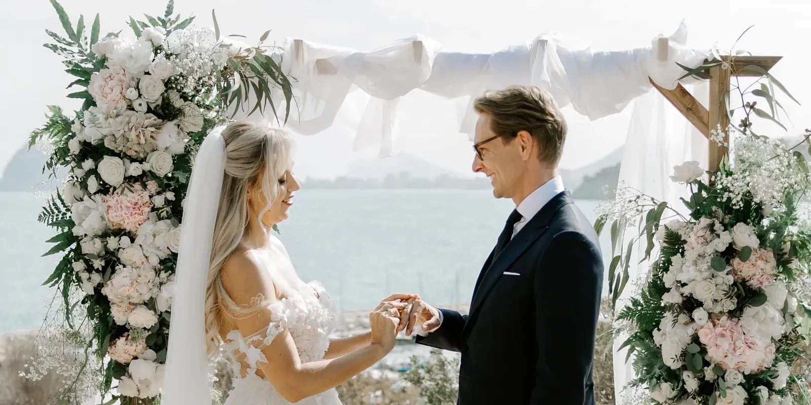 Bride and groom exchanging rings under a flower arch, with the Mediterranean behind